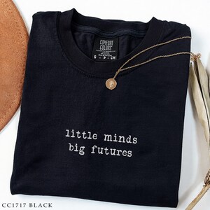 May include: Black t-shirt with the text "little minds big futures" in white. A gold-colored necklace with a round pendant rests on the shirt. The shirt is labeled "Comfort Colors" and the style is CC1717 BLACK.