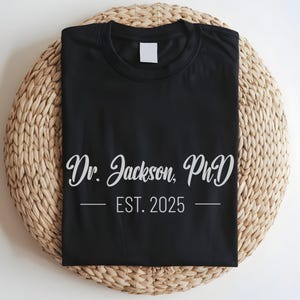 May include: Black t-shirt with white text that reads "Dr. Jackson, PhD - EST. 2025"