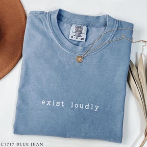 May include: A blue jean colored t-shirt with the words "exist loudly" in white script. A gold necklace with a pendant rests on the shirt. The shirt is folded and has a tag that says "Comfort Colors".