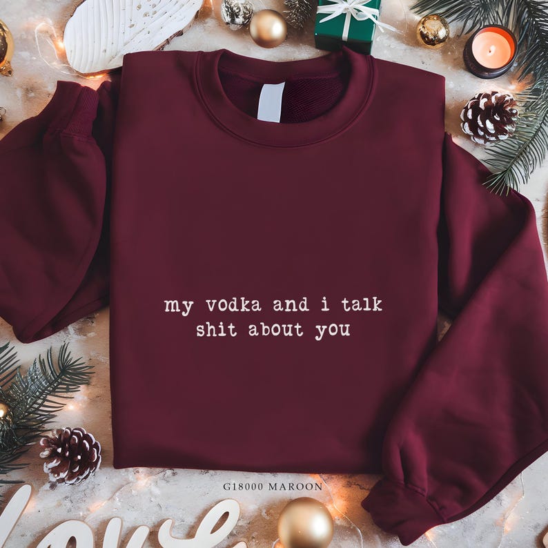 May include: A maroon sweatshirt with the white text "my vodka and i talk shit about you". The long-sleeved, crew-neck top has the product code "G18000 MAROON" printed at the bottom. The sweatshirt is displayed on a festive background with Christmas decorations.