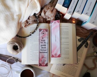 May include: A pink and white bookmark with the quote "It made her the greatest threat he'd ever encountered" from the book "Crown of Midnight". The bookmark is laying on an open book with a cup of coffee, a feather pen, and a sword.