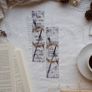 Puede incluir: Dos marcadores decorativos con fondo blanco y texto negro. El marcador superior presenta una cita del libro "The Crown of Gilded Bones" de Jennifer Armentrout. La cita dice: "You will bow before your queen or you will bleed before her. It is your choice.". El marcador inferior presenta el título del libro y el nombre del autor.