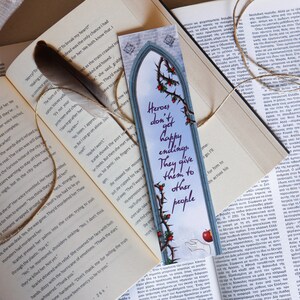 Puede incluir: Un marcap&aacute;ginas azul y gris con un dise&ntilde;o de arco g&oacute;tico y una vid espinosa con rosas rojas. El marcap&aacute;ginas presenta una cita en texto morado: "Los h&eacute;roes no tienen finales felices. Se los dan a los dem&aacute;s."
