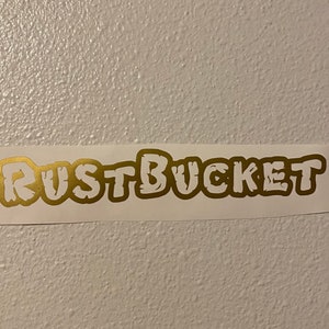 May include: Gold vinyl sticker with the text "RUST BUCKET" in a stylised font.