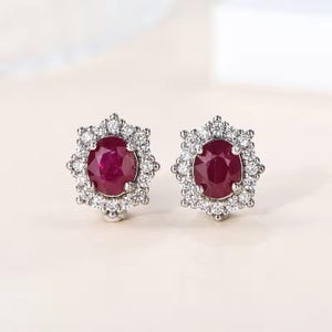 May include: A pair of stud earrings featuring oval-cut deep red rubies at the center. Each ruby is surrounded by a halo of small, clear, round-cut white stones set in a silver-toned metal frame, creating a floral-like shape for each earring.