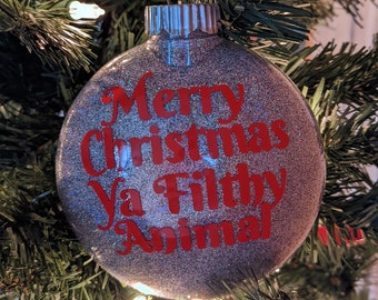 Op de afbeelding: Een zilveren glitter kerstbal met de tekst "Merry Christmas Ya Filthy Animal" in het rood. De bal hangt aan een rood-wit gestreepte draad, tegen de achtergrond van een kerstboom.