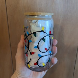 May include: A glass jar with a bamboo lid, decorated with a black outline of Christmas lights in red, yellow, blue, and green. The jar is filled with white tissue paper.