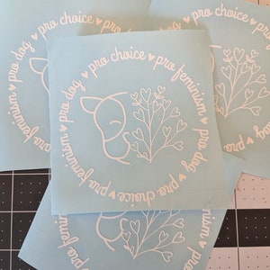 May include: Three blue vinyl stickers with white text and a white outline of a dog with a heart-shaped flower. The text on the stickers reads "pro dog pro choice pro feminism".