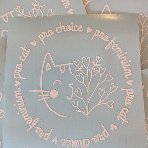 May include: A light blue sticker with a white outline of a cat with a heart-shaped nose and a plant with hearts on the branches. The text around the cat and plant reads "pro choice pro feminism pro cat".