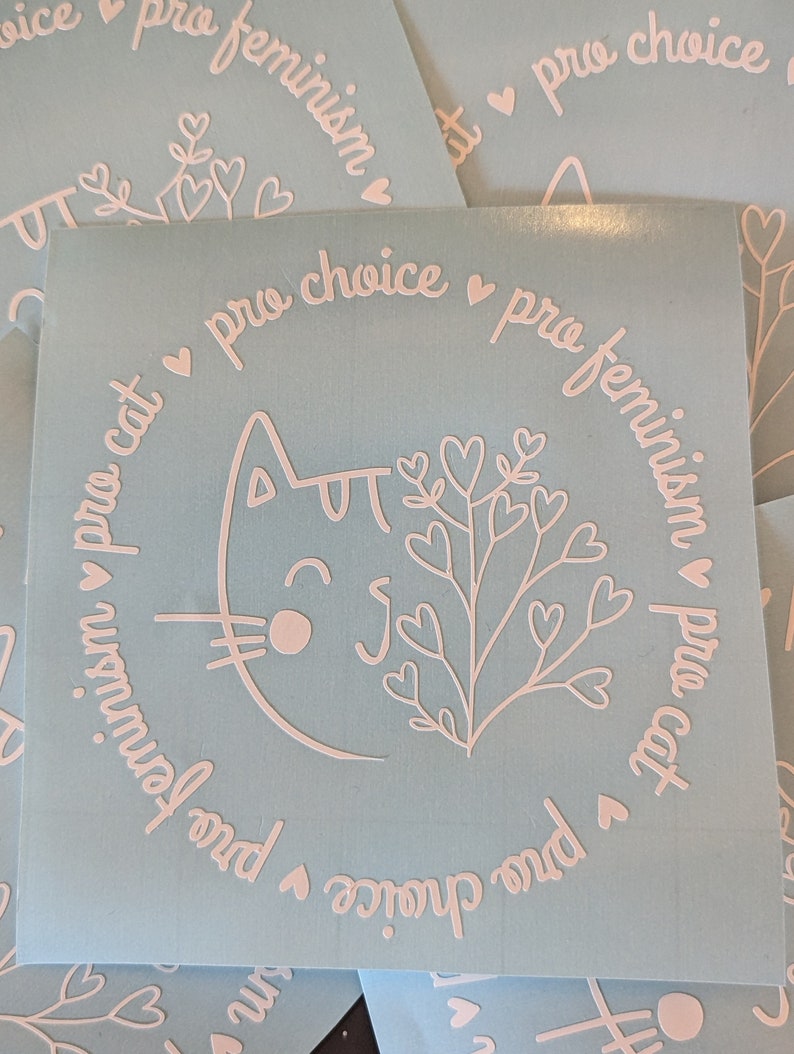 May include: A light blue sticker with a white outline of a cat with a heart-shaped nose and a plant with hearts on the branches. The text around the cat and plant reads "pro choice pro feminism pro cat".