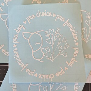 May include: A light blue sticker with a white outline of a dog sniffing a flower with hearts. The sticker has the text "pro dog pro choice pro feminism" in a circle around the dog and flower.