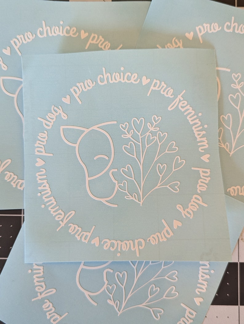 May include: A light blue sticker with a white outline of a dog sniffing a flower with hearts. The sticker has the text "pro dog pro choice pro feminism" in a circle around the dog and flower.