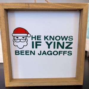 May include: Wooden framed sign with the text "HE KNOWS IF YINZ BEEN JAGOFS" in green letters. A cartoon Santa Claus with a red hat and white beard is in the upper left corner. The sign is hanging by a natural fiber rope.