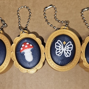 May include: Four gold-colored pendants with silver chains. Each pendant has a dark blue background and a different design: a sword, a mushroom, a butterfly, and a book. The pendants have a decorative, scalloped edge.