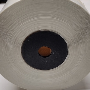 May include: A roll of white thermal paper for a label printer. The paper is rolled around a black core with a small hole in the center.