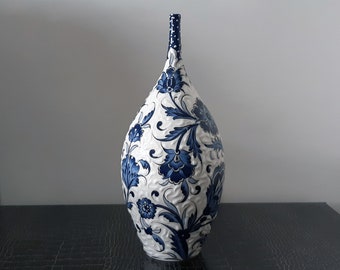 May include: A tall, white ceramic vase with a slender neck and bulbous body. The vase is adorned with a repeating pattern of blue floral designs. The neck features a dotted pattern, and the overall design is embossed.