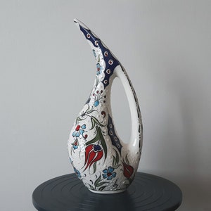 May include: A white ceramic jug with a long, curved spout and a handle. The jug is decorated with a colourful floral pattern in shades of blue, red, and green.