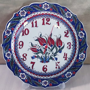Könnte beinhalten: Eine weiße Keramikuhr mit einem blauen und roten Blumendesign. Das Zifferblatt der Uhr zeigt römische Ziffern und ein Blumenmotiv. Die Uhr steht auf einem weißen Ständer.