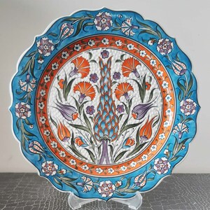 Pode incluir: Um prato de cer&acirc;mica azul e branco com um design floral. O prato apresenta um motivo central de uma flor estilizada com folhas laranja, azuis e verdes. A borda do prato &eacute; decorada com um padr&atilde;o repetitivo de flores menores.