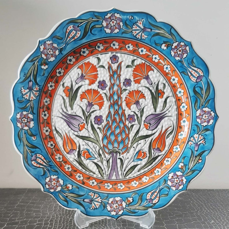 Pode incluir: Um prato de cer&acirc;mica azul e branco com um design floral. O prato apresenta um motivo central de uma flor estilizada com folhas laranja, azuis e verdes. A borda do prato &eacute; decorada com um padr&atilde;o repetitivo de flores menores.