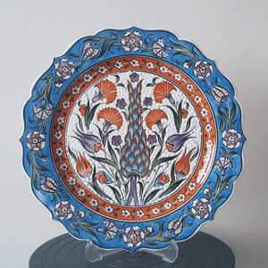 Pode incluir: Um prato de cer&acirc;mica decorativo com uma borda azul e branca e um desenho central de flores laranja e roxas sobre um fundo branco.