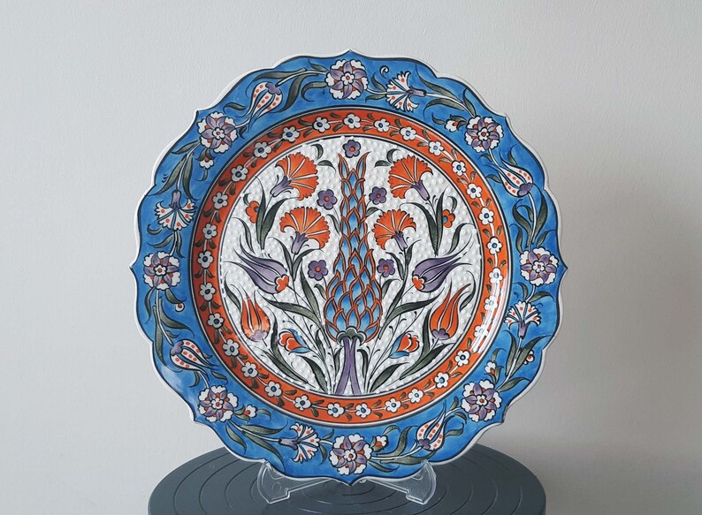 Pode incluir: Um prato de cer&acirc;mica decorativo com uma borda azul e branca e um desenho central de flores laranja e roxas sobre um fundo branco.