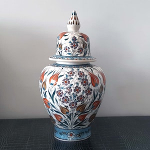 May include: A white ceramic vase with a lid, decorated with a floral pattern in shades of blue, orange, and green. The vase has a bulbous body and a narrow neck.