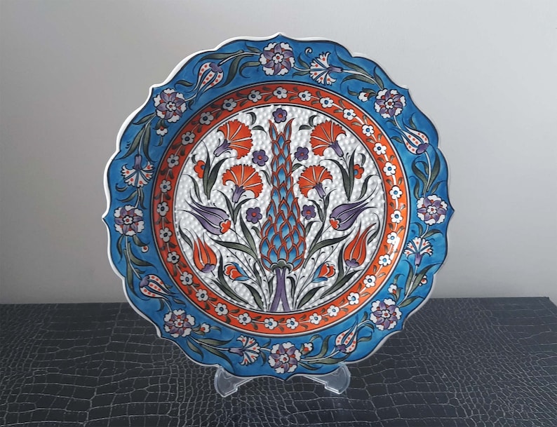 Pode incluir: Um prato de cer&acirc;mica branca com um design floral azul e laranja. O prato tem uma borda festonada e &eacute; decorado com um motivo central de uma flor estilizada com um centro azul e laranja. O prato est&aacute; sobre um suporte preto.