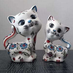 May include: Two white ceramic cat figurines with blue, red, and green floral designs. The cats are sitting upright with their heads tilted up.