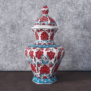 May include: A white ceramic jar with a lid, decorated with red and blue floral patterns. The jar has a hexagonal base and a rounded top.