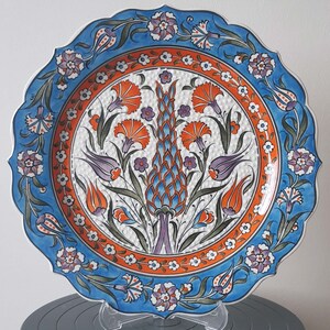 Pode incluir: Um prato redondo de cer&acirc;mica com um design floral azul e branco. O prato apresenta um motivo central de uma flor estilizada com acentos laranja, azul e verde. O prato tem uma borda festonada e &eacute; decorado com um padr&atilde;o repetitivo de pequenas flores brancas.