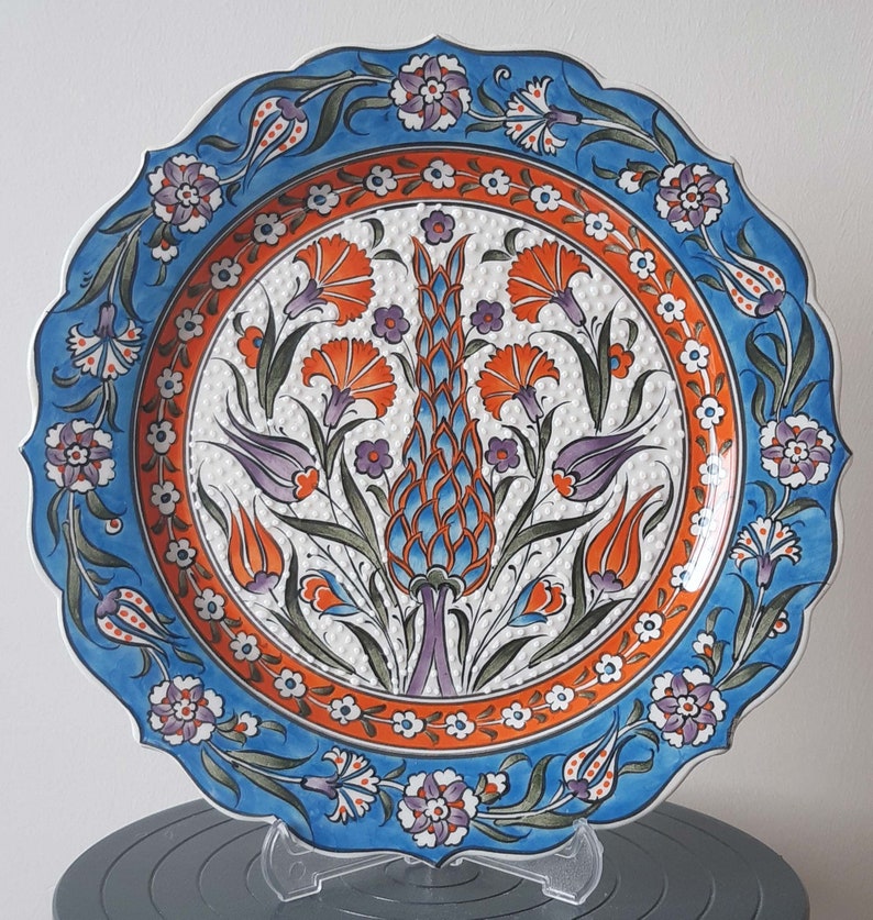 Pode incluir: Um prato redondo de cer&acirc;mica com um design floral azul e branco. O prato apresenta um motivo central de uma flor estilizada com acentos laranja, azul e verde. O prato tem uma borda festonada e &eacute; decorado com um padr&atilde;o repetitivo de pequenas flores brancas.
