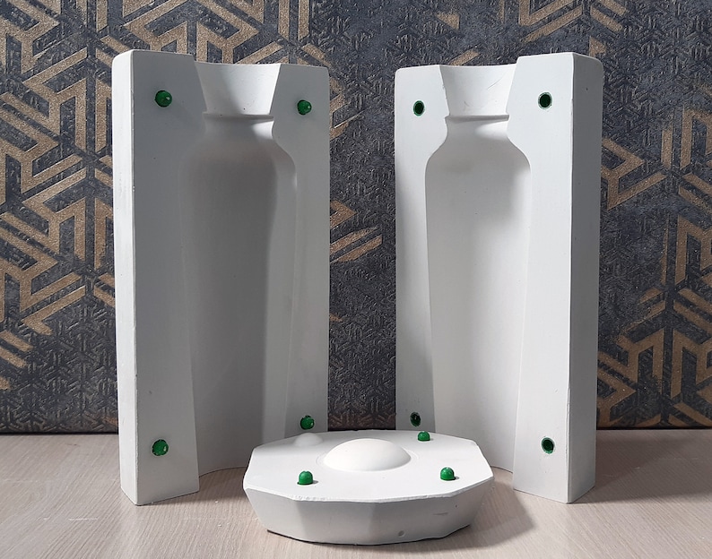 May include: Two white plaster molds for making bottles. The molds are open on the top and bottom, and have a round base. The molds are sitting on a light brown surface.