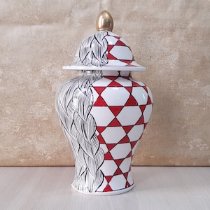 Può includere: Un vaso di zenzero in ceramica bianca con un motivo geometrico rosso e bianco su un lato e un motivo astratto bianco e nero sull'altro. Il vaso ha un coperchio dorato.
