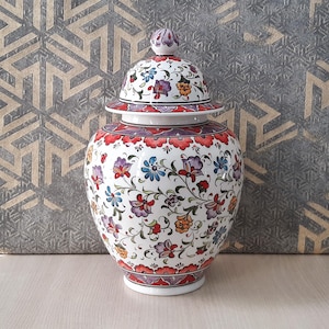May include: A white ceramic jar with a lid, decorated with a colorful floral pattern in shades of red, blue, and yellow. The jar has a round body and a slightly flared top.