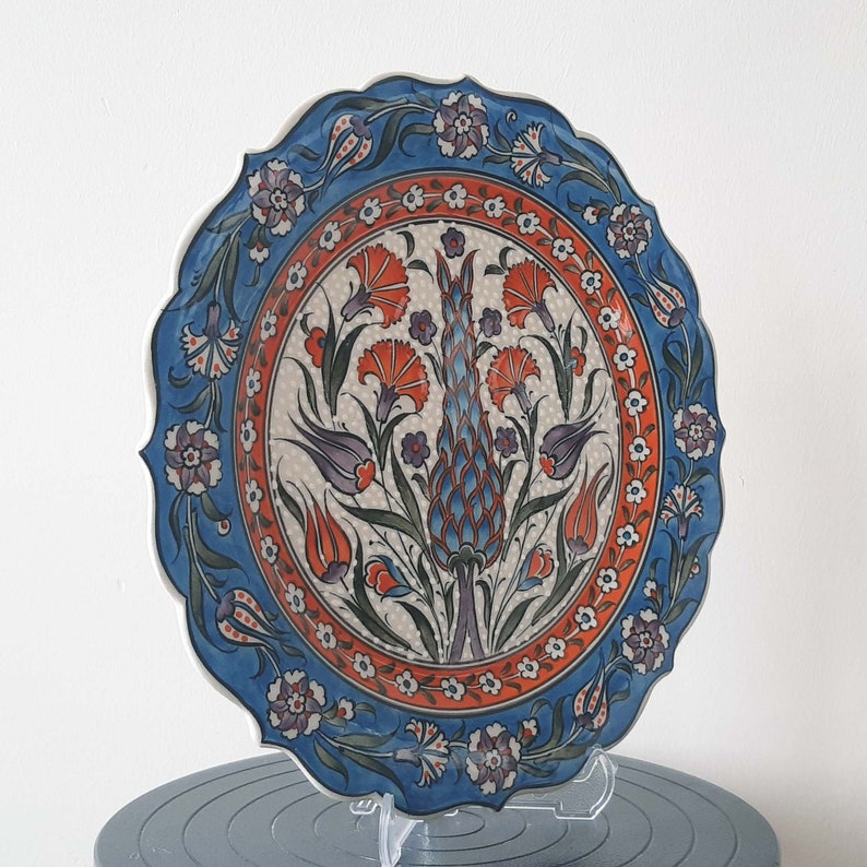 Pode incluir: Um prato de cer&acirc;mica azul e branco com um design floral. O prato apresenta um motivo central de uma flor estilizada com acentos vermelhos, laranja e roxos. O prato tem uma borda festonada e &eacute; decorado com um padr&atilde;o repetitivo de flores e folhas.