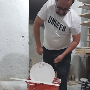 Pode incluir: Uma pessoa vestindo uma camiseta branca com a palavra "UNSEEN" impressa nela est&aacute; segurando um prato de cer&acirc;mica branco sobre um balde vermelho. O prato est&aacute; suspenso por um gancho de metal.