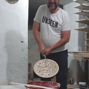 Pode incluir: Um homem vestindo uma camiseta branca com a palavra "UNSEEN" impressa nela, segura um prato redondo de cer&acirc;mica com um design floral em dourado e branco. O prato est&aacute; pendurado em um gancho de metal.