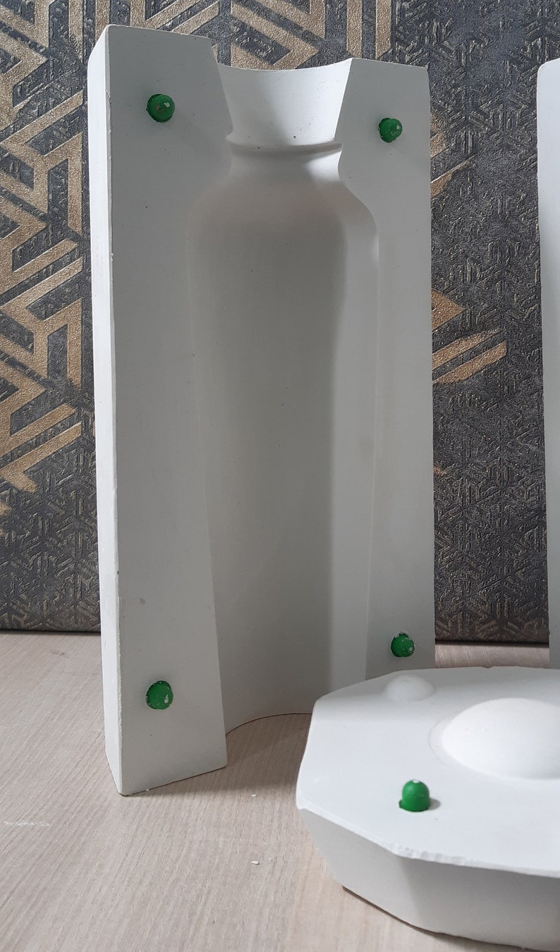 May include: White plaster mold for making a bottle or vase. The mold has a smooth, rounded interior and a textured exterior. There are five green dots on the mold.