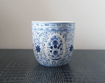 Puede incluir: Un macetero de cerámica blanca con un patrón floral azul y blanco. El macetero tiene una forma redondeada y un borde ancho.