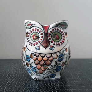 May include: White ceramic owl figurine with colorful floral and geometric patterns. The owl has large, round eyes with brown pupils and a brown beak.