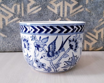 Puede incluir: Un macetero de cerámica blanca con un diseño floral azul y blanco que presenta un pájaro. El macetero tiene un patrón de chevron alrededor del borde superior.