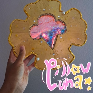 Può includere: Un oggetto decorativo dorato a forma di quadrifoglio con un design centrale iridescente. Il quadrifoglio è delineato con una corda dorata e adornato con piccoli cristalli e perle. Il testo "Pillow Luna Patterns" è visibile.