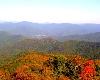 May include: A scenic landscape of rolling green mountains under a clear blue sky. The foreground features trees with vibrant red and orange foliage, suggesting an autumn season. The image captures a panoramic view of the natural environment.