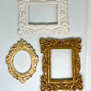 May include: Three ornate picture frames in different shapes and colors. A white rectangular frame is at the top, a gold oval frame is in the lower left, and a gold rectangular frame is in the lower right. All frames have detailed, decorative designs.