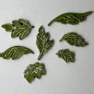 May include: Seven green ceramic leaf-shaped tiles with detailed veins and textures. The tiles are arranged in a semi-circle on a white background.