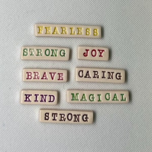 May include: A set of eight ceramic tiles with positive words written on them. The words are "Fearless", "Strong", "Joy", "Brave", "Caring", "Kind", "Magical", and "Strong". The tiles are in various colors, including yellow, green, red, purple, and black.