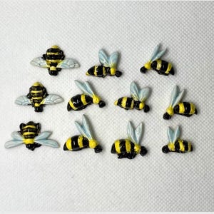 May include: Twelve ceramic bee figurines with black and yellow stripes and white wings. The bees are arranged in a random pattern on a white surface.
