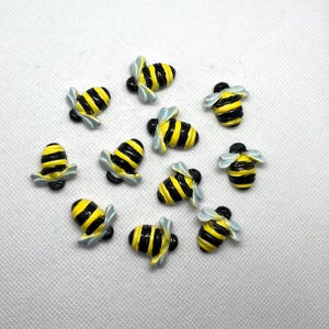 May include: A collection of small, decorative bee-shaped embellishments. Each bee features a yellow and black striped body, with light blue wings. The bees are arranged on a white background.
