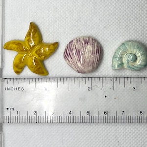 Puede incluir: Tres adornos de cer&aacute;mica con tem&aacute;tica marina: una estrella de mar amarilla, una concha p&uacute;rpura y blanca y una concha azul y blanca. Los adornos miden aproximadamente 2,5 cm.